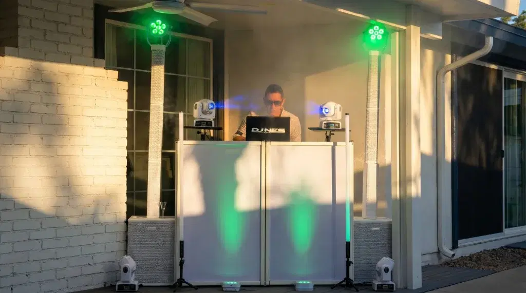 DJ NES behind a white booth on an outdoor patio with column speakers, green and blue stage lighting, and light haze