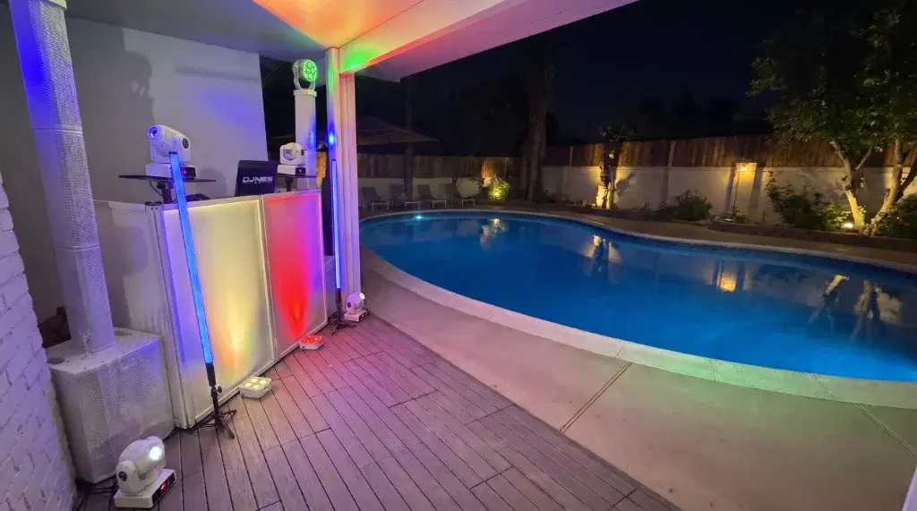 Mobile DJ booth with column speakers and colorful LED lighting beside a glowing pool at a nighttime backyard party