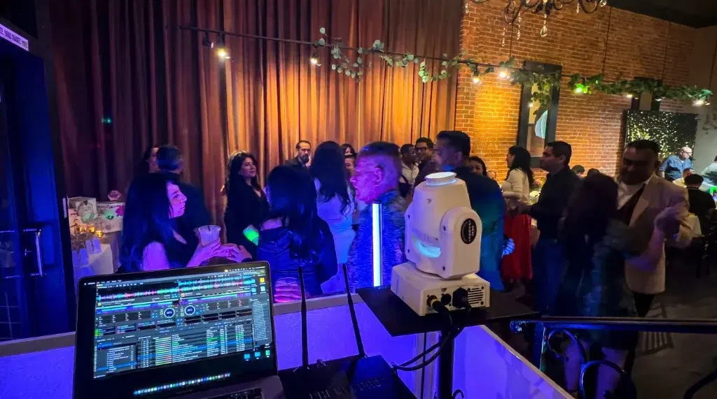 View from the DJ booth with laptop and moving-head light overlooking a packed dance floor under warm string lights and blue uplighting