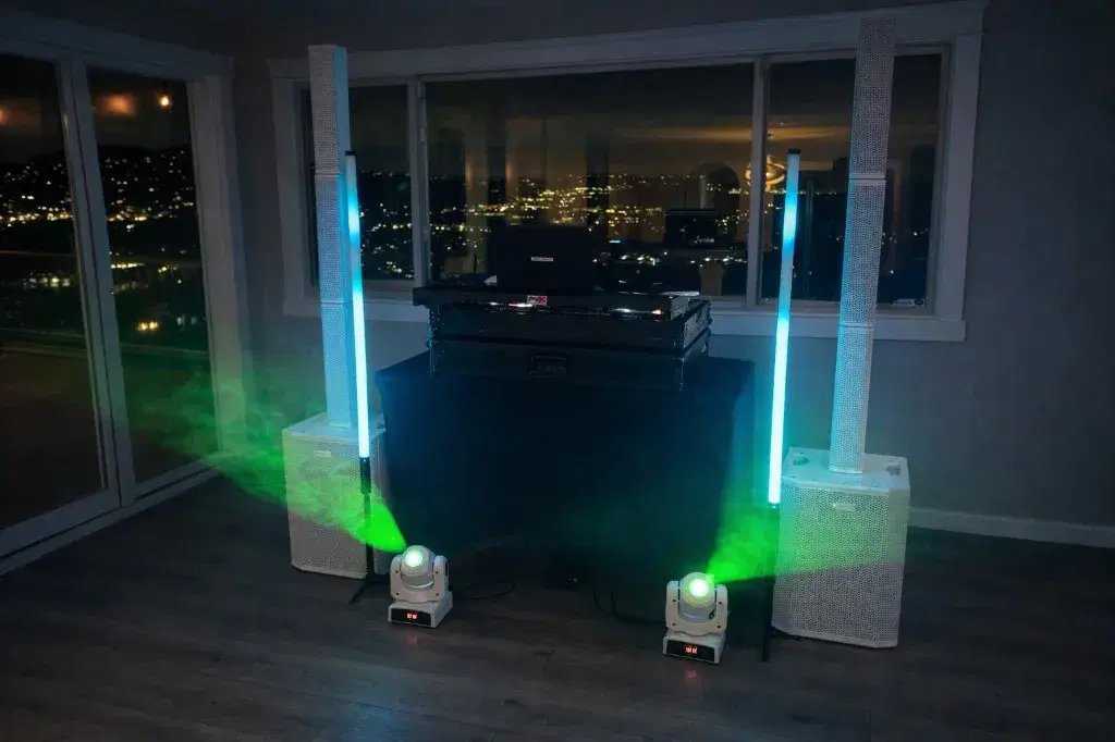 Professional mobile DJ setup with white column speakers, cyan LED tubes, and green spotlights overlooking a city at night
